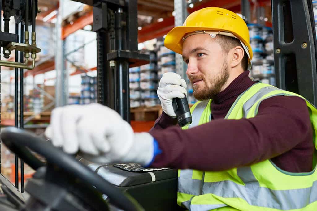 Waist up portrait of young man sitting in forklift and using walkie-talkie while moving goods in warehouse, copy space
