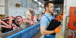 Bicycle factory, worker with a walkie-talkie poses at bike assembly line. Male mechanic in uniform installs cycle parts in workshop, industrial manufacturing