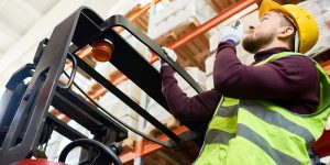 Low angle portrait of young man sitting in forklift and using walkie-talkie while moving goods in warehouse, copy space